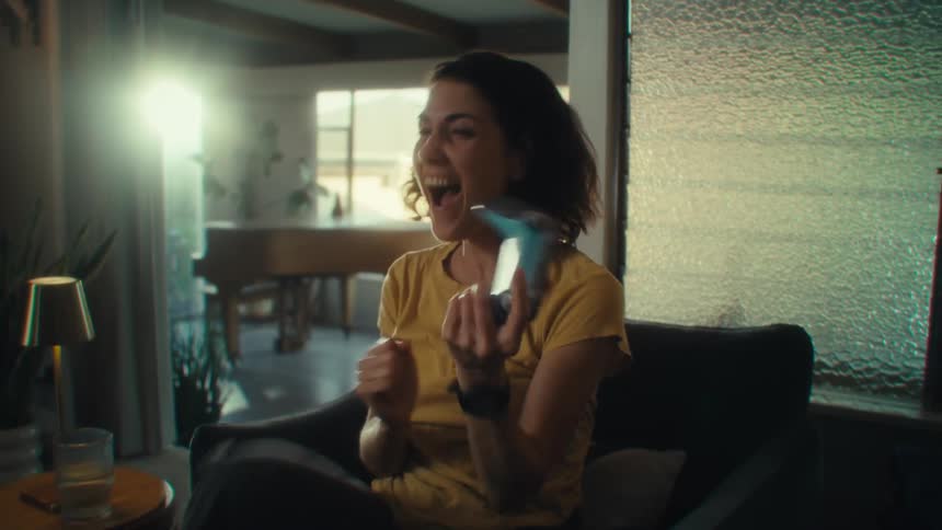 A young woman sits excitedly on her couch holding a gaming controller and playing a video game moving around with enthusiasm