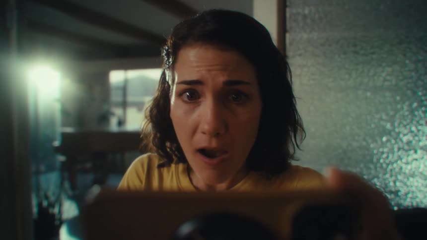A woman with dark hair wearing a yellow shirt is looking down at a smartphone she holds in both hands as her expression changes from concerned to shocked and excited