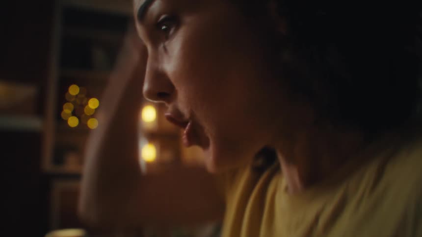 A close view shows a woman with dark hair and a light yellow shirt looking off to her right a big smile on her face and a blurry living room in the background