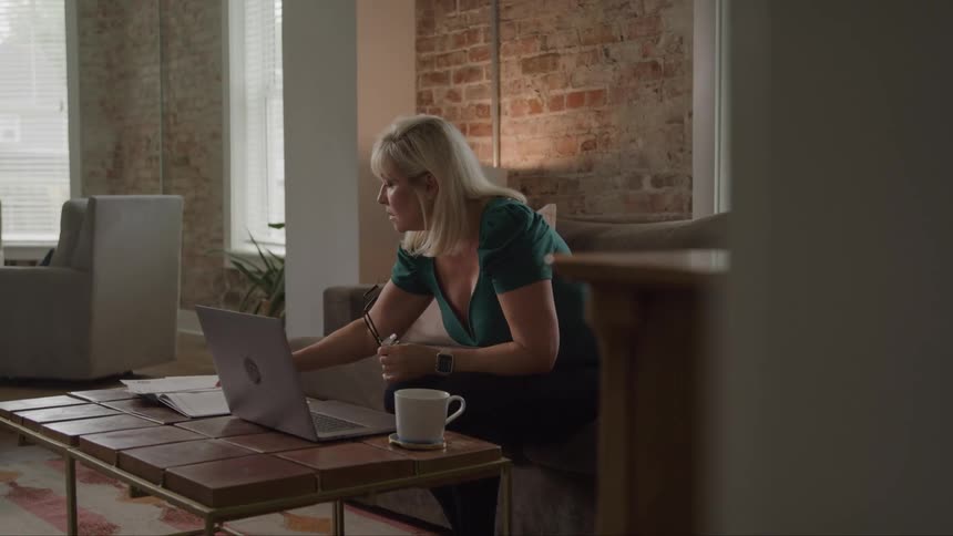 A woman with long blond hair is sitting on a couch in a living room holding her glasses in her hands while looking down at documents next to a laptop that is open on the coffee table in front of her