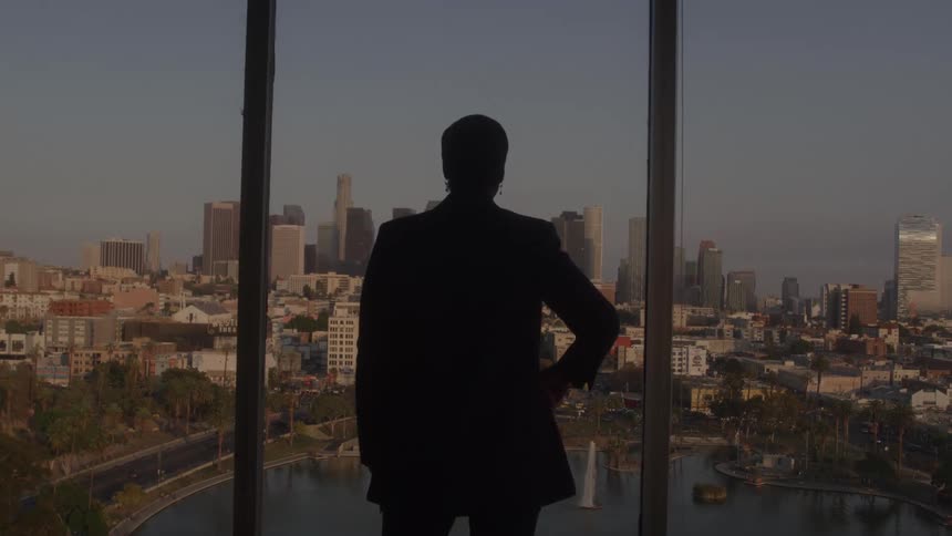 A woman stands in front of a window inside an office building at dawn gazing at the cityscape