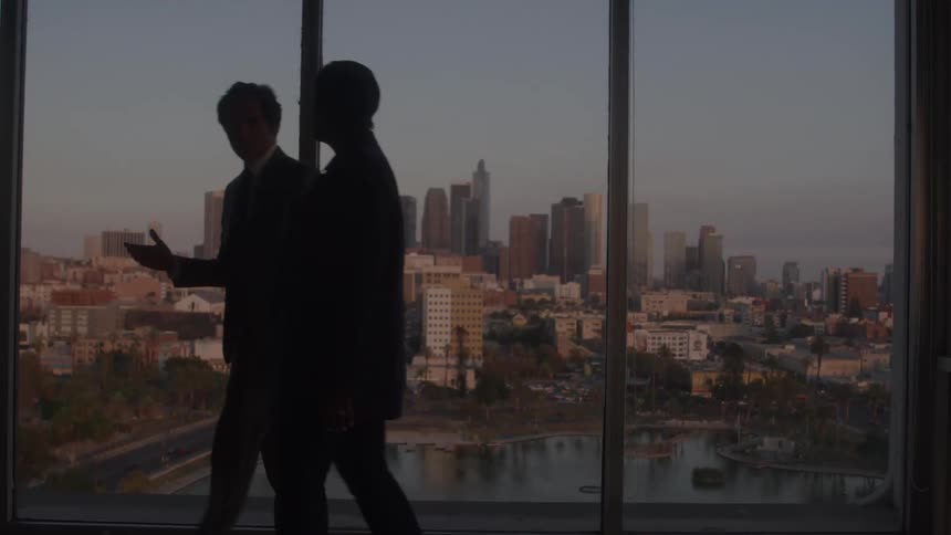 The video shows two business people standing in silhouette against a large window overlooking a city they appear to be talking to one another and gesturing towards the cityscape