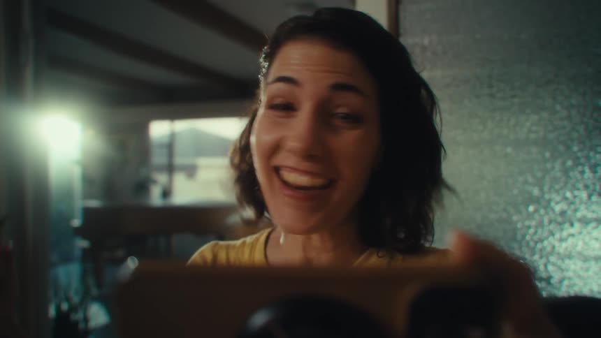 A woman with dark hair wearing a yellow shirt is looking down at a smartphone she holds in both hands with an excited expression then she smiles and laughs