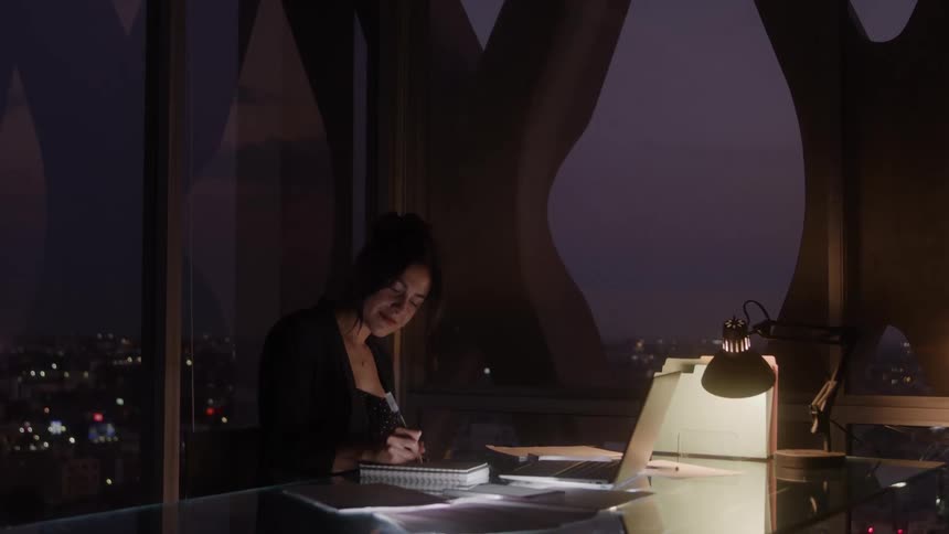 A woman with long dark hair is sitting at a desk with a laptop and a pile of notebooks as she writes in one of the notebooks with a pen while a lamp provides light and a city skyline is visible through the large window behind her