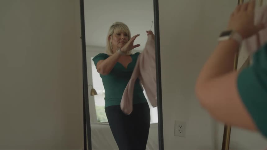 A woman with blond hair wearing a green shirt and black pants is standing in front of a mirror and looking at a pale pink blouse she holds on a hanger