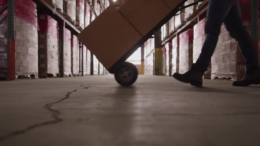 The video shows the interior of a large warehouse with tall shelves full of boxes the camera is low to the ground and moves forward down an aisle toward a bright light source at the end