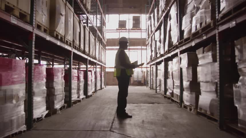 A middle aged black male worker walks while inventory in a warehouse during daytime