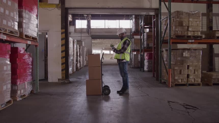 A warehouse worker wearing a white hard hat and a yellow safety vest is standing in a warehouse aisle next to a hand truck with a box on it while he looks down adjusting a glove in his hands