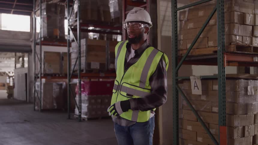 A male warehouse worker wearing a hard hat safety glasses and a high visibility vest winces and holds his lower back as if in pain surrounded by tall shelves stacked with boxes