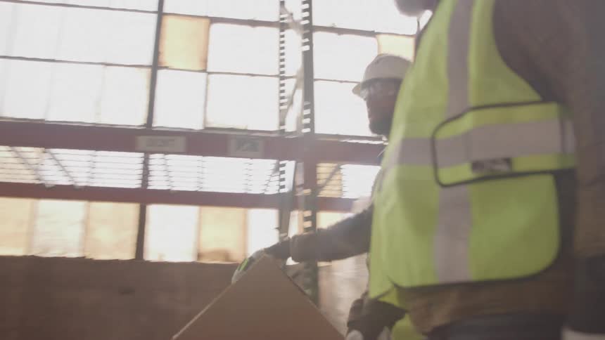 Three workers wearing reflective vests and hard hats in a warehouse one pushing a cart with boxes while another takes notes on a clipboard