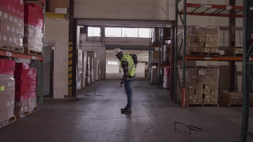 Worker wearing a reflective vest and hard hat standing in the middle of a warehouse looking at the floor