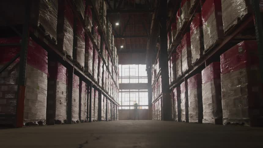 A view of a warehouse aisle shows workers in safety vests working between tall shelvesfull of boxes