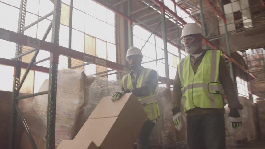 Two male coworkers both wearing hard hats and yellow safety vests are carrying a large cardboard box down a warehouse aisle while a third coworker walks past them