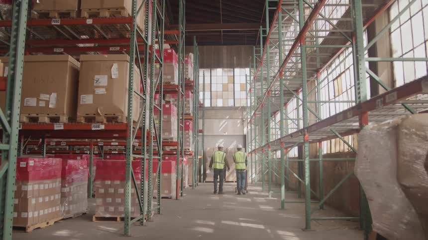 Two men wearing hard hats and green vests push a trolley down a warehouse aisle filled with shipping boxes at midday