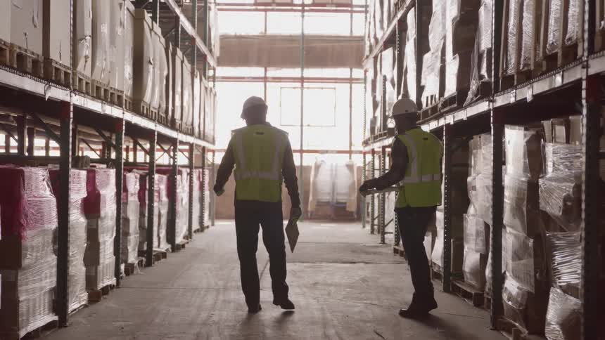 Silhouettes of two workers wearing reflective vests and hard hats walking between warehouse shelves