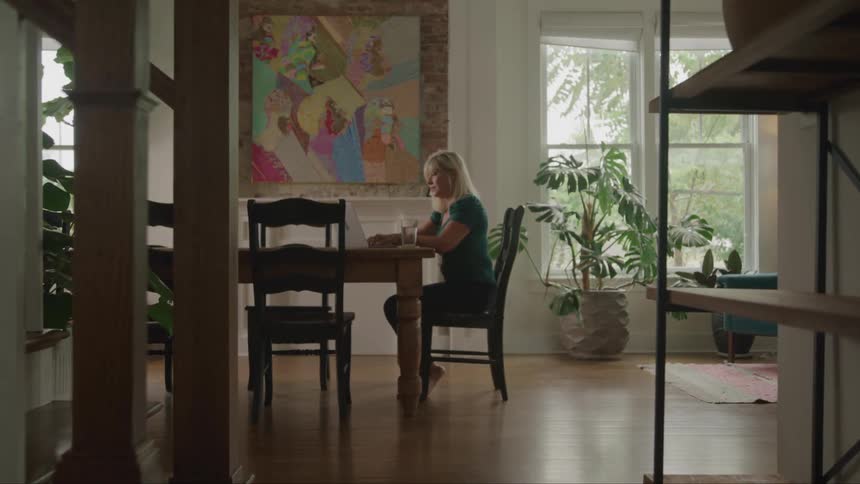 A woman with blond hair wearing a dark green shirt and dark pants is sitting at a wooden dining table in a room with a large colorful painting on the wall behind her as she types on a laptop