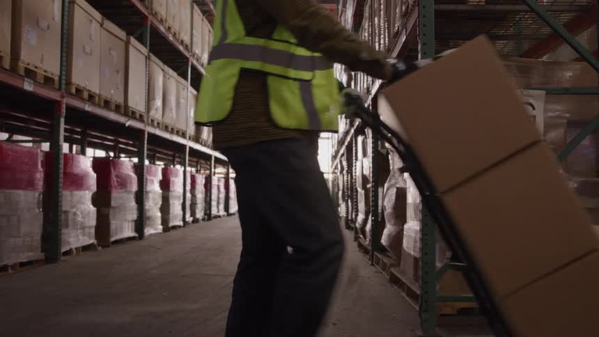 A man wearing a hard hat and a yellow safety vest is walking down a warehouse aisle lined with tall shelves of boxes while pushing a hand truck with a stack of three boxes on it