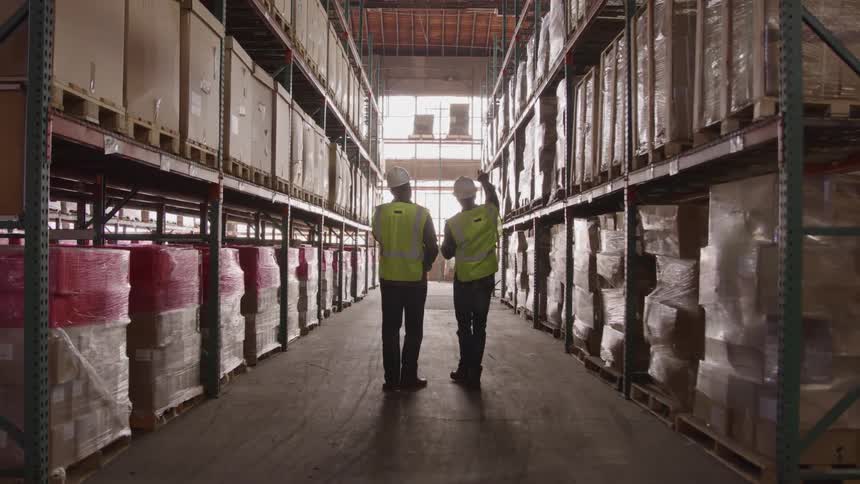 Two male workers walk inside a distribution shipping warehouse wearing hard hats and green vests with rows of shipping boxes stacked on shelves
