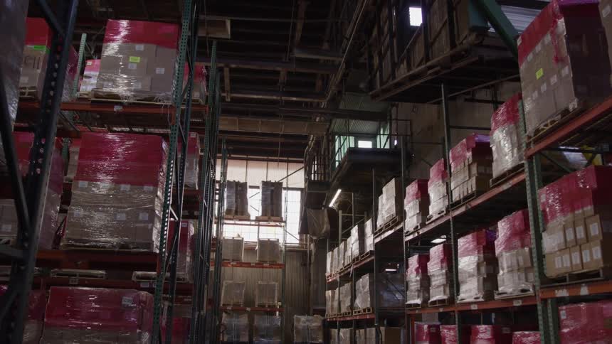 A long view shows the inside of a warehouse with a high ceiling and tall shelves full of boxes as the camera slowly moves forward down the main aisle