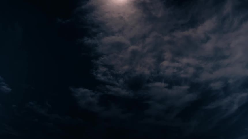 A low angle view of a night sky shows the moon shining brightly through dark swirling clouds