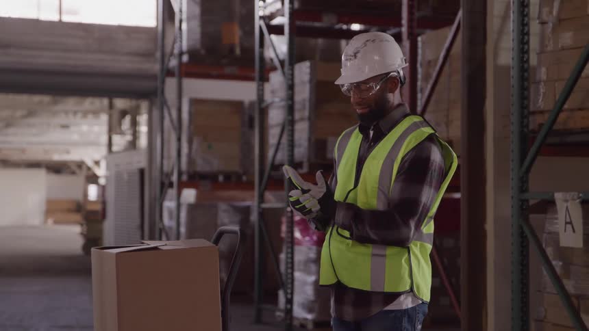 Worker wearing a reflective vest and hard hat looking at a device in his hands near a box in a warehouse