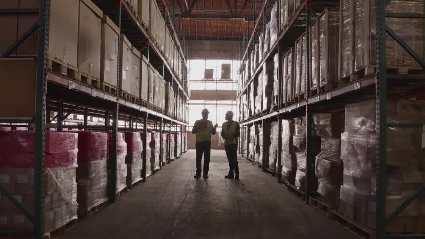 Two men both wearing hard hats and safety vests are standing in a warehouse aisle with tall shelves full of boxes as they talk and another worker walks past them