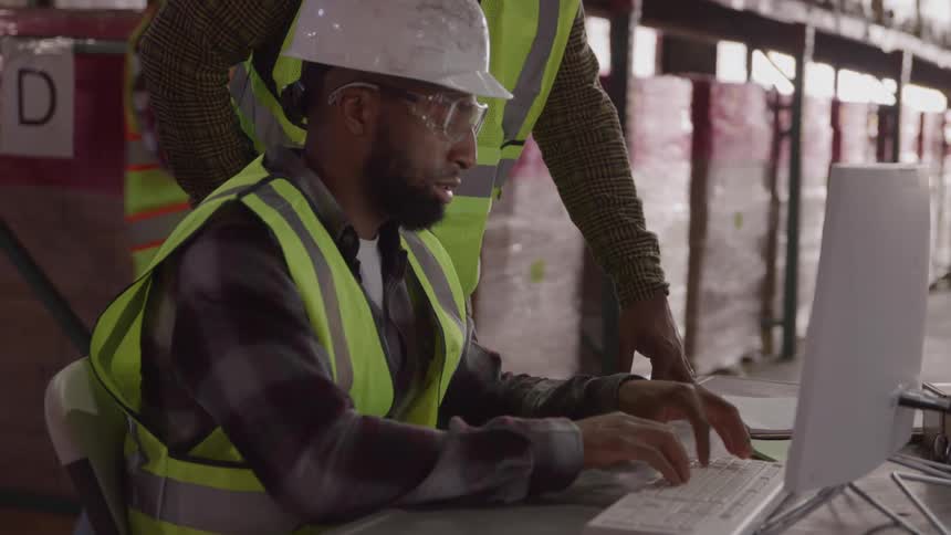A warehouse worker with a black beard wearing a white hard hat a plaid shirt a yellow safety vest and clear safety glasses is sitting at a desk and typing on a computer keyboard in a warehouse while a coworker stands next to him