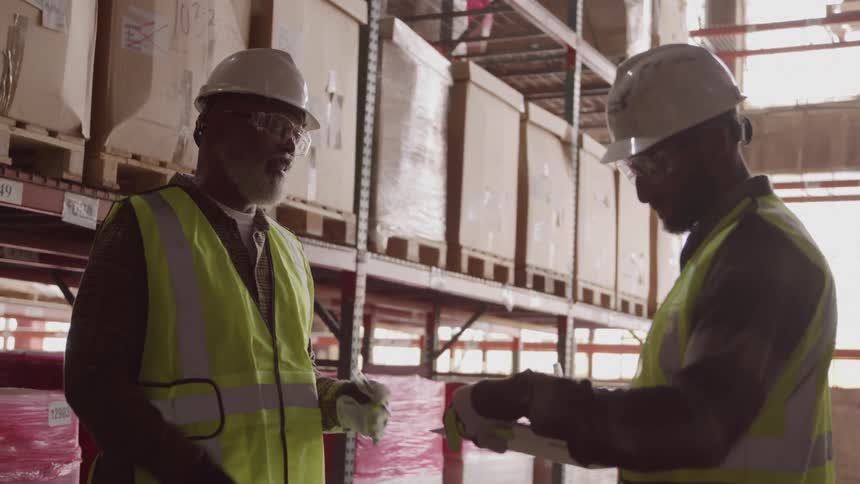 Two workers wearing reflective vests and hard hats talking inside a warehouse