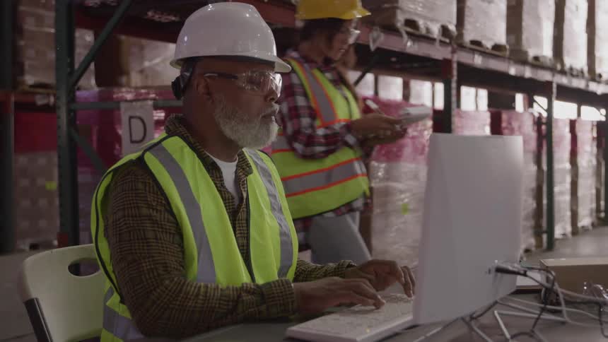 A man with a white beard wearing a white hard hat a plaid shirt a yellow safety vest and clear safety glasses is sitting at a desk and typing on a computer keyboard in a warehouse aisle while another worker walks past him in the background