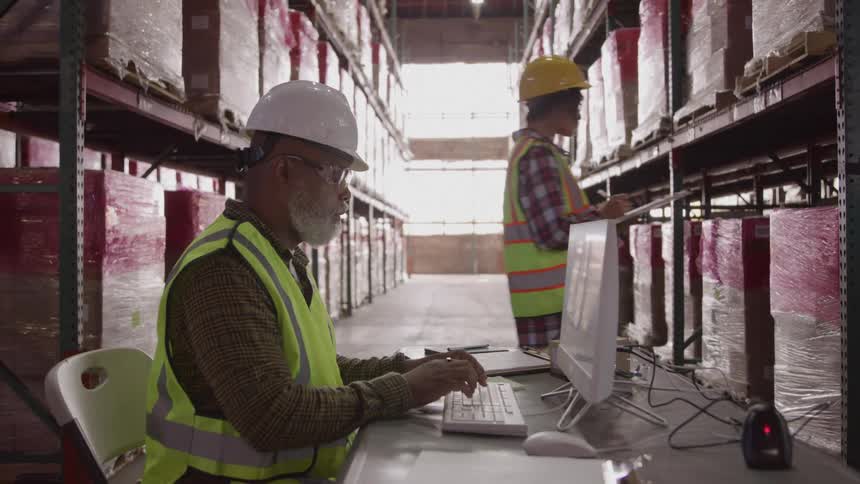 Worker wearing a reflective vest and hard hat typing on a computer in a warehouse while another inspects shelves in the background