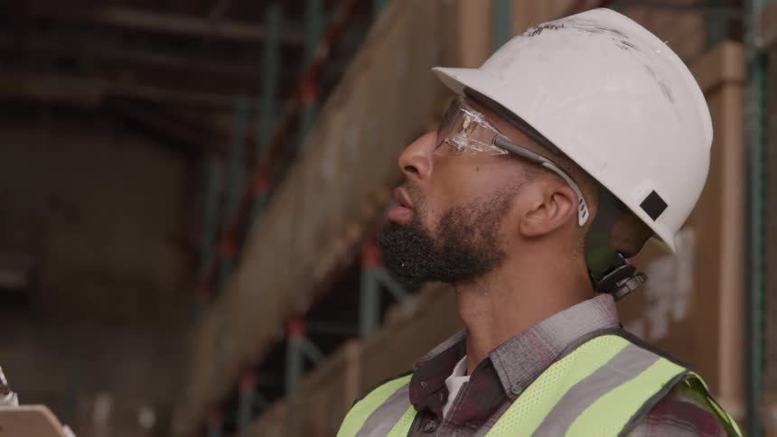 A man with a black beard wearing a white hard hat a plaid shirt a yellow safety vest and clear safety glasses is standing in a warehouse and looking up from a clipboard he is holding in his hands