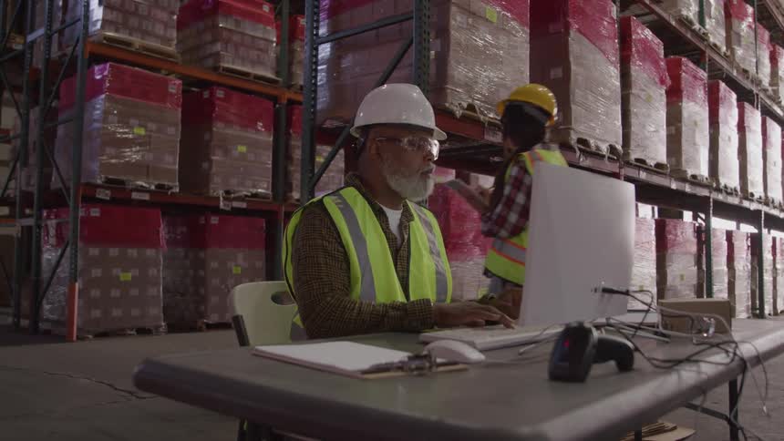 A man with a white beard wearing a white hard hat a plaid shirt a yellow safety vest and clear safety glasses is sitting at a desk and typing on a computer keyboard in a warehouse while another worker walks past him