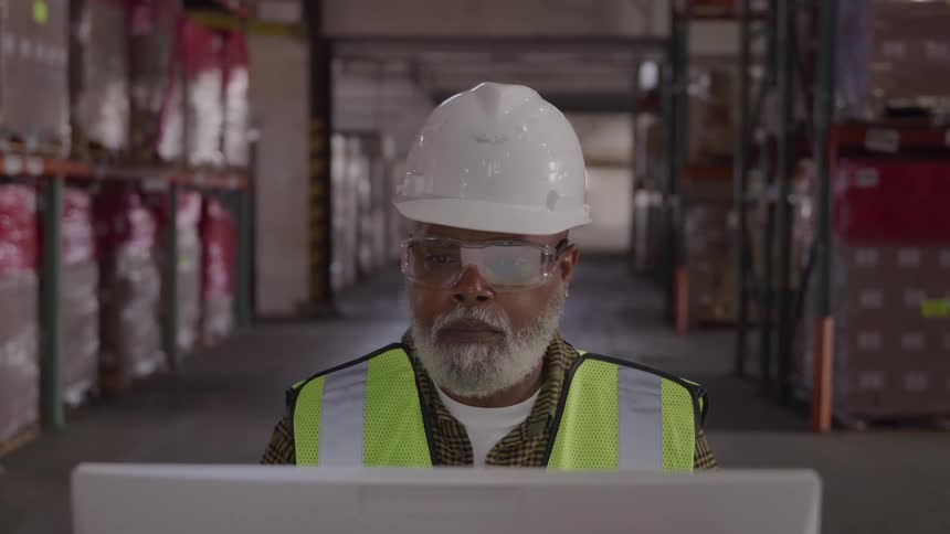 Worker wearing a reflective vest and helmet looking at a computer in the warehouse