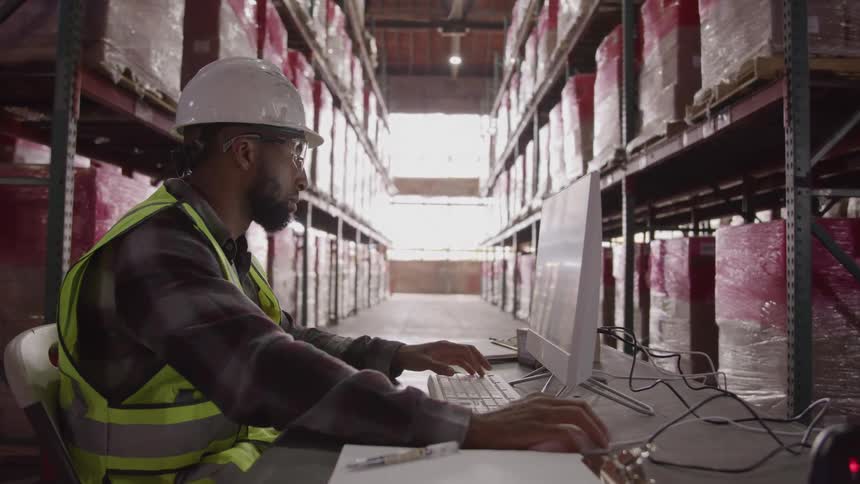 Worker wearing a reflective vest and hard hat using a computer in a warehouse aisle