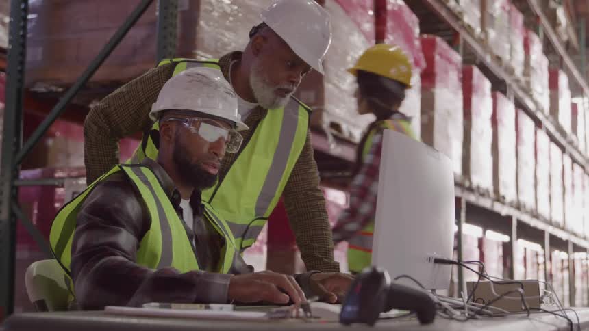 Two men in hard hats working in front of a computer in a storage facility