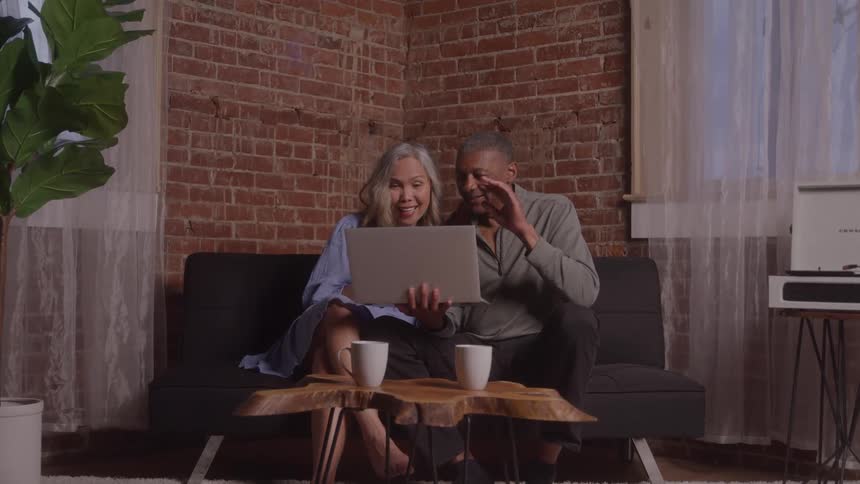 An elderly couple wave while on a video call with a laptop as they are seated on a couch in a room indoors during daytime