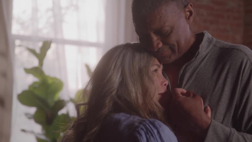 A couple is dancing in a room with a brick wall and a large window as the woman rests her head on the mans chest while he holds her close and they sway together