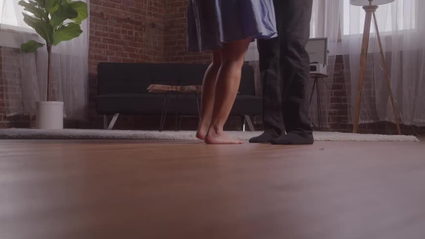 A view of two people from the waist down shows a woman standing barefoot and a man wearing black shoes as they dance close together on a wooden floor