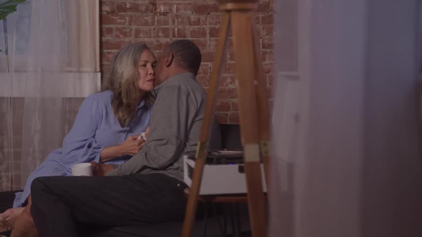 A man and a woman sitting each holding a glass leaning forward and kissing