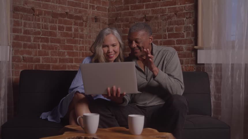 An elderly man sits beside a woman on a couch holding a laptop during a video call and waving at the caller with two coffee mugs on the table in front of them