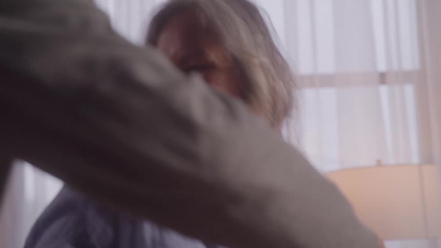 A woman with long wavy gray and brown hair smiles brightly as she dances with a man whose back is turned to the camera the couple moves together in a warm home setting
