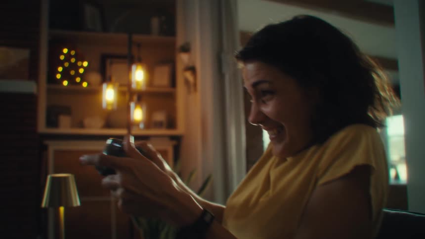 A woman with dark hair wearing a yellow shirt is sitting in a chair and playing a video game on a controller as she reacts with an excited and laughing expression