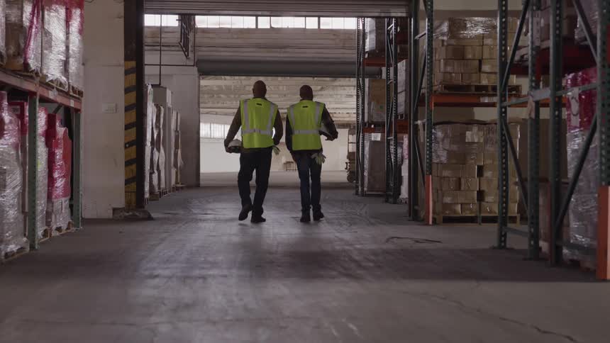 Two workers in reflective vests walking down a warehouse aisle while another person passes in the foreground