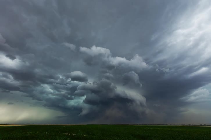 A tornado is visible in the distance on a grassland as dark clouds cover the sky at midday