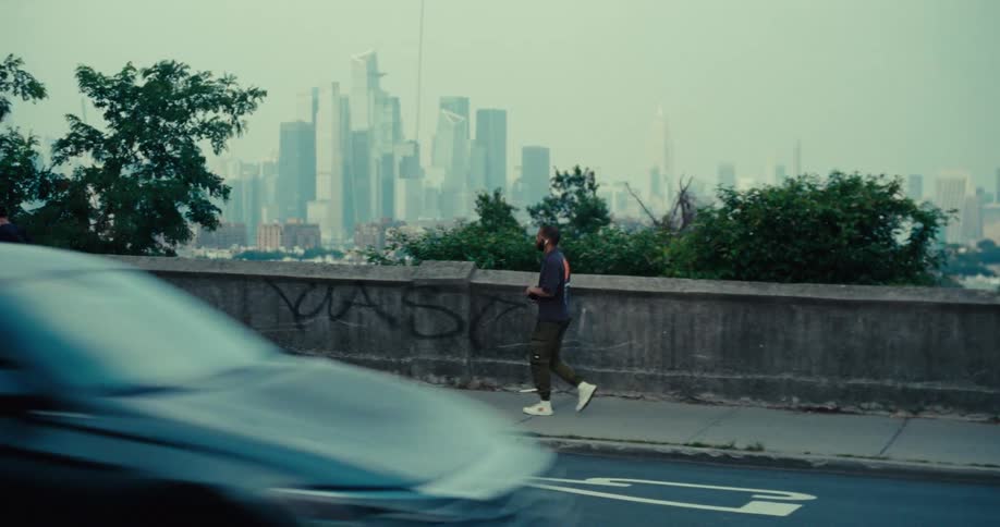 Two men walk along a sidewalk by a concrete wall one with a dog against a hazy city skyline backdrop