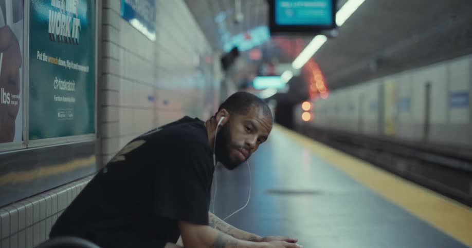 A man with earbuds sits on a subway platform looking around while waiting in the urban underground station with a tunnel and other commuters in view