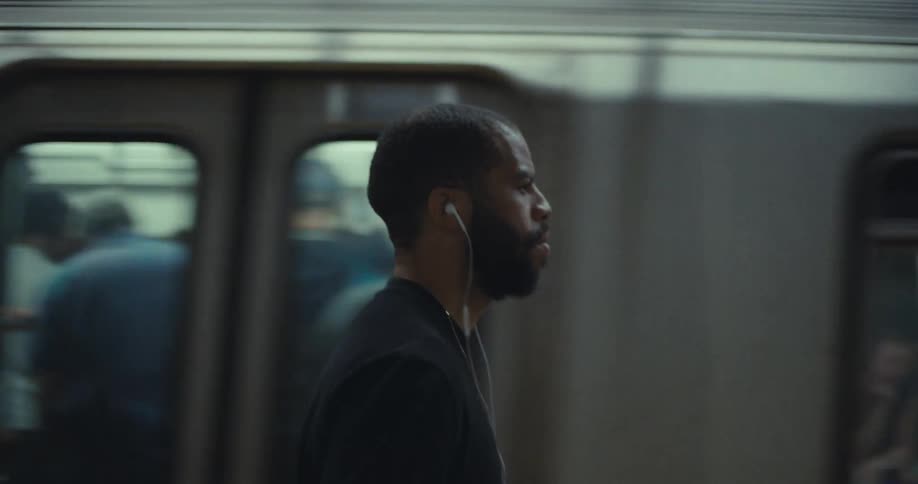 A medium shot shows a man standing on a train platform with a train passing behind him he is looking ahead and appears to be listening to music with wired earphones