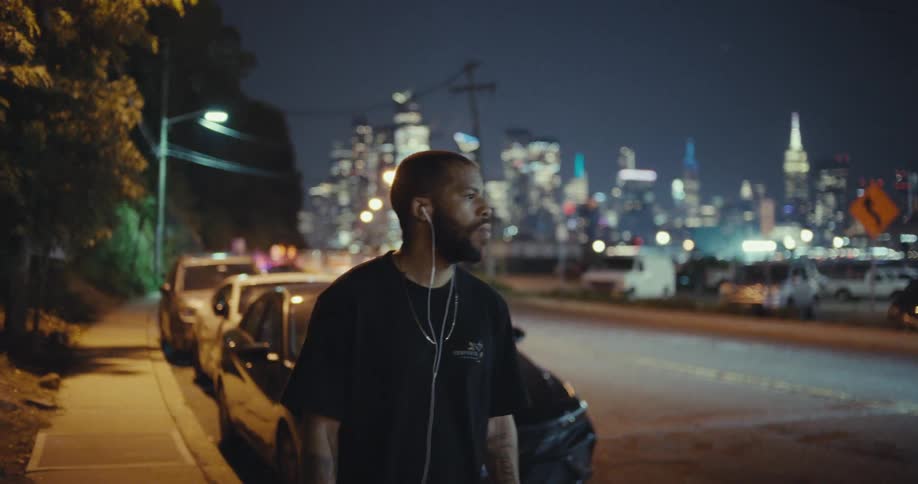 The video shows a man standing on a sidewalk at night listening to music with headphones the background features a blurred cityscape with bright lights and a row of cars parked on the street
