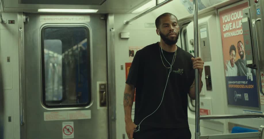 A man wearing a black tshirt stands holding a pole inside a train car during a ride placing his phone in his pocket with wired earbuds attached as he enjoys his music