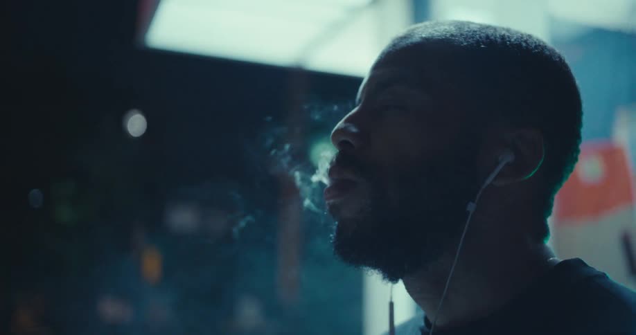 At night under the bright lights of a bus stop a man lights a handrolled cigarette takes a deep puff and then exhales a thick cloud of smoke as he tilts his head back to look up